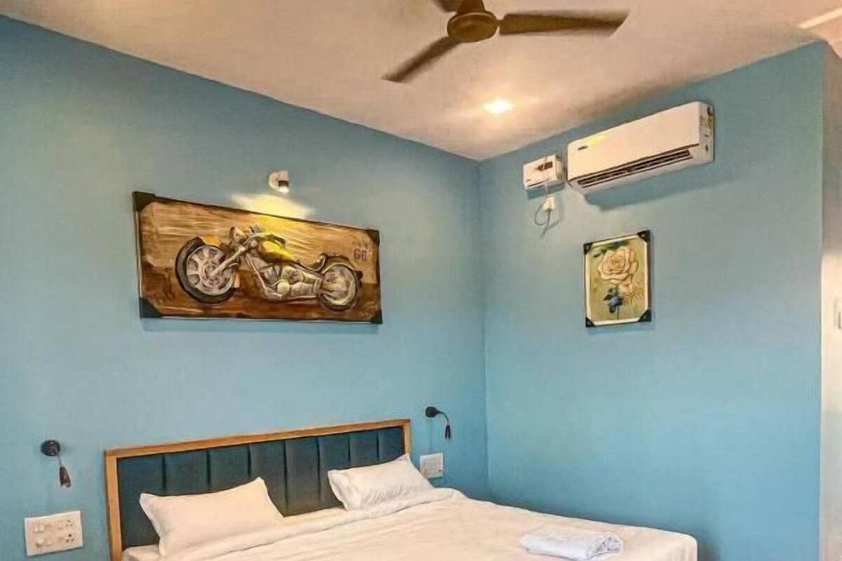 A bedroom with a wooden bed, a ceiling fan, an air conditioner, and a wall-mounted motorcycle painting.