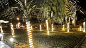 Ein Außenbereich bei Nacht mit Palmen und beleuchteten Laternen, einem Schachbrettmuster-Weg und einem grünen Mülleimer.