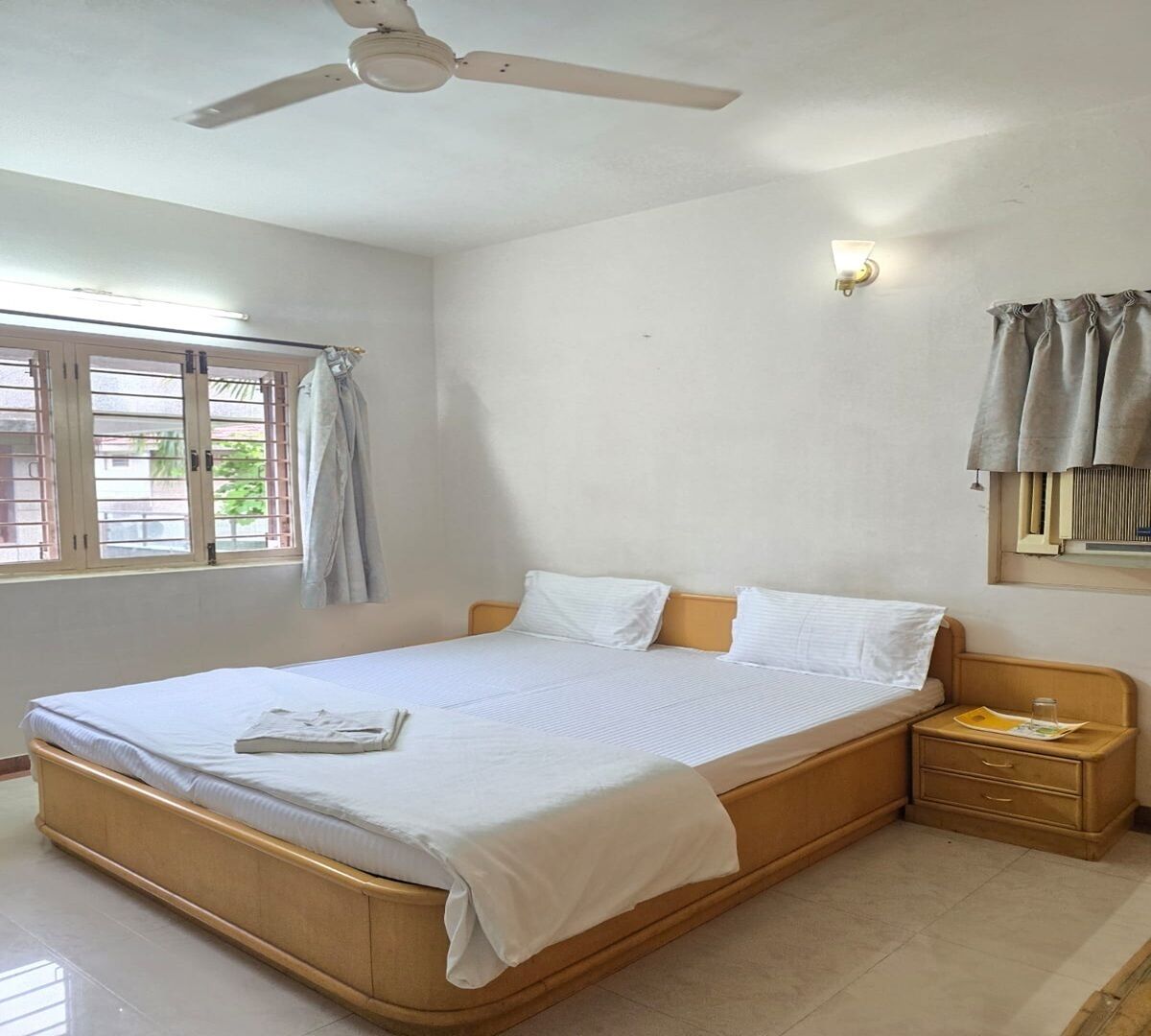 Un dormitorio con cama, mesita de noche, ventilador de techo, ventana con cortinas y una lámpara fijada a la pared.