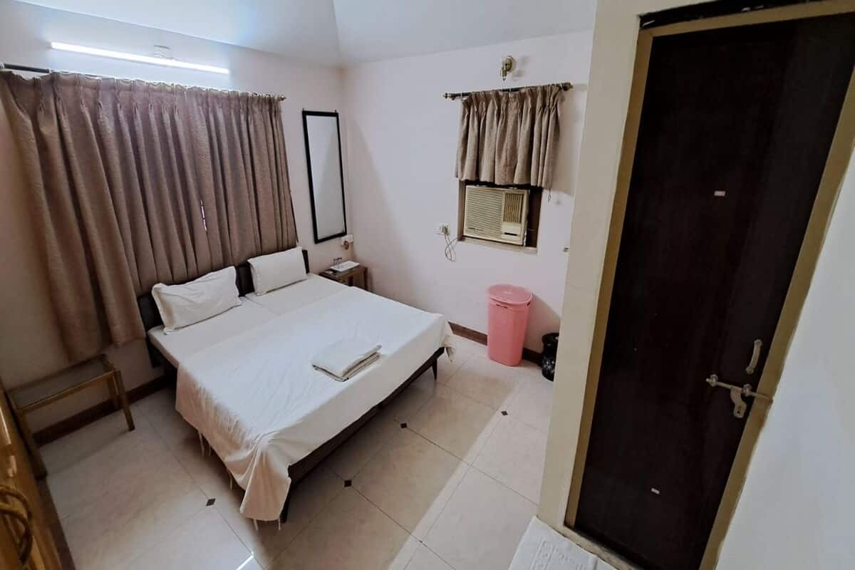A bedroom with a bed, a nightstand, a mirror, a window with curtains, a door, and a small window with an air conditioner.