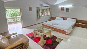 A hotel room with a bed, a ceiling fan, a glass table, a sofa, and a view of greenery outside. - Elis Hospitality Service Parth Bungalow (Ahmedabad)
