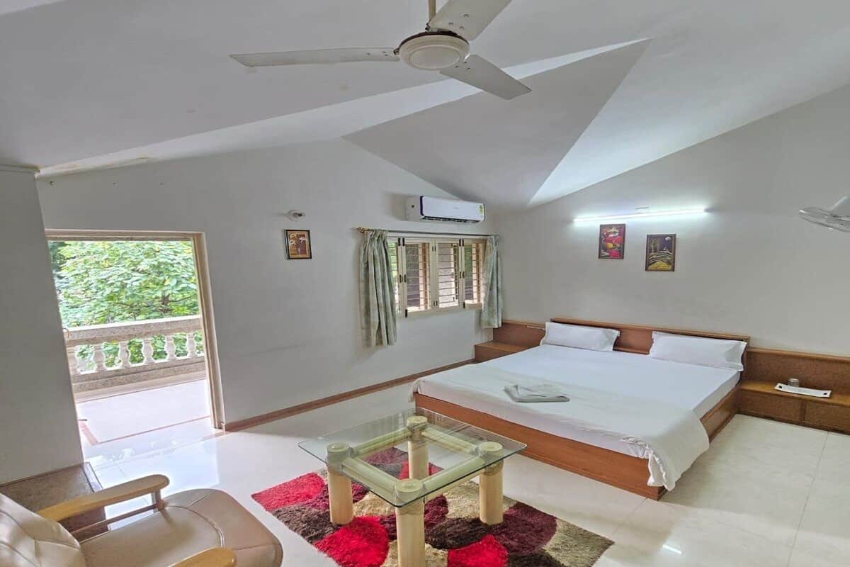A hotel room with a bed, a ceiling fan, a glass table, a sofa, and a view of greenery outside.