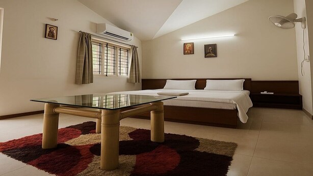 A modern bedroom with a glass-top table, a bed with white linens, a red and beige rug, and a ceiling fan. - Elis Hospitality Service Parth Bungalow (Ahmedabad)