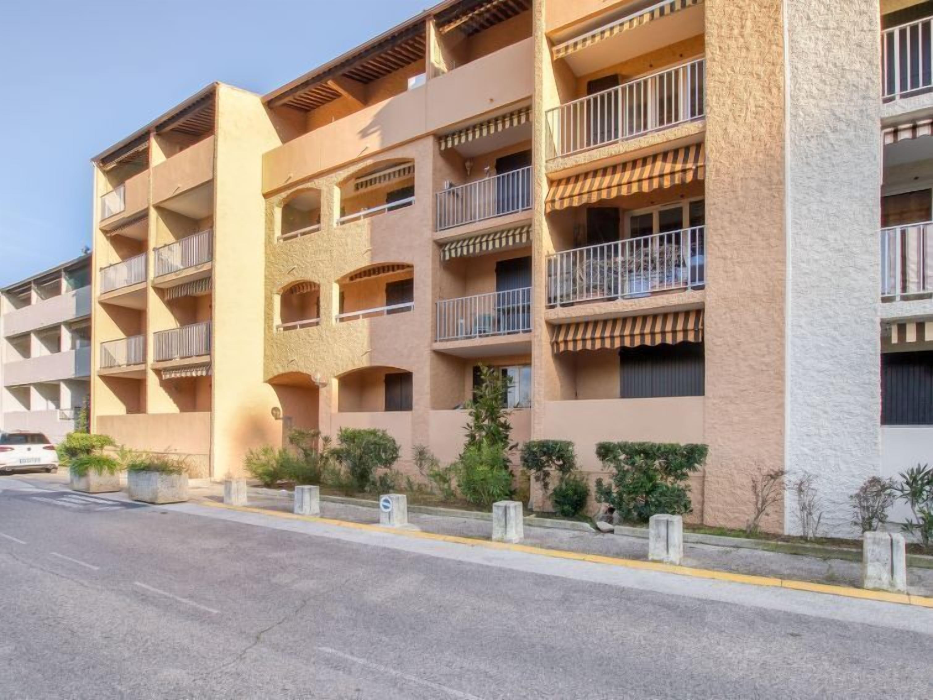 Un immeuble résidentiel à plusieurs étages, doté de balcons, donnant sur une rue et bordé de petits arbustes le long du trottoir.