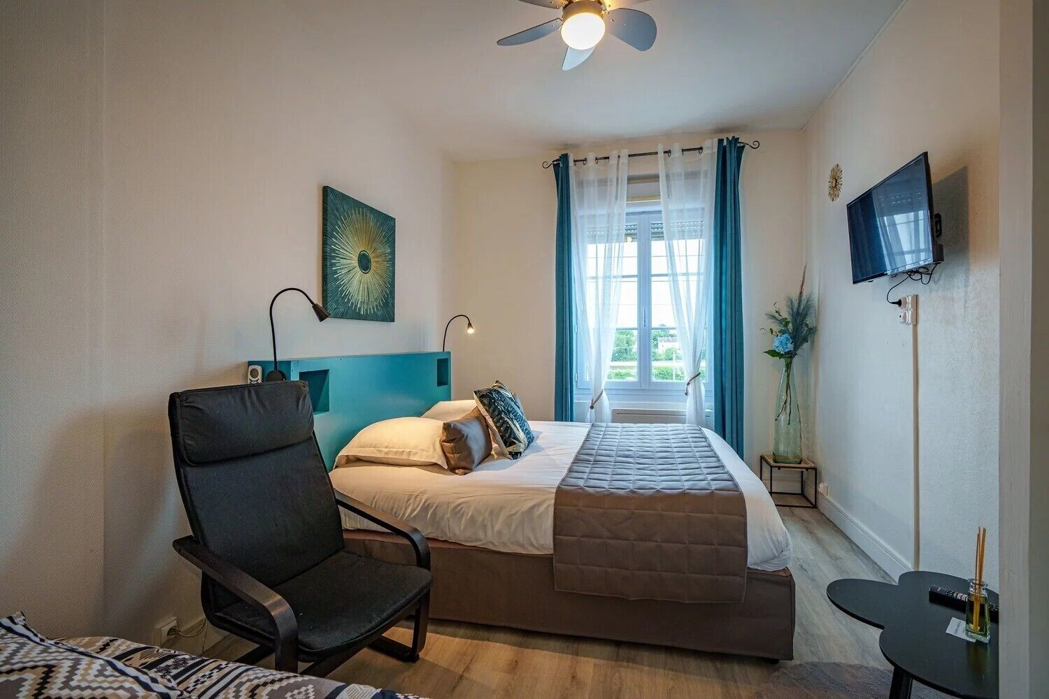 A bedroom with a bed, a chair, a TV, a ceiling fan, and a window with curtains.