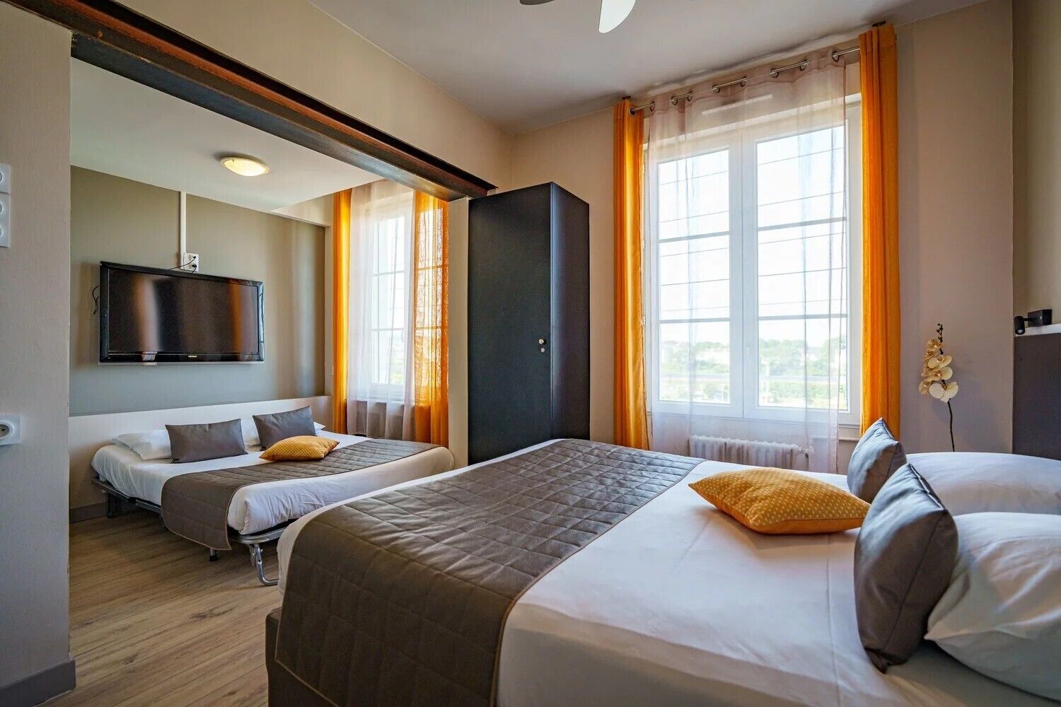 A hotel room with two beds, a flat-screen TV, a large window with curtains, and a view of greenery outside.