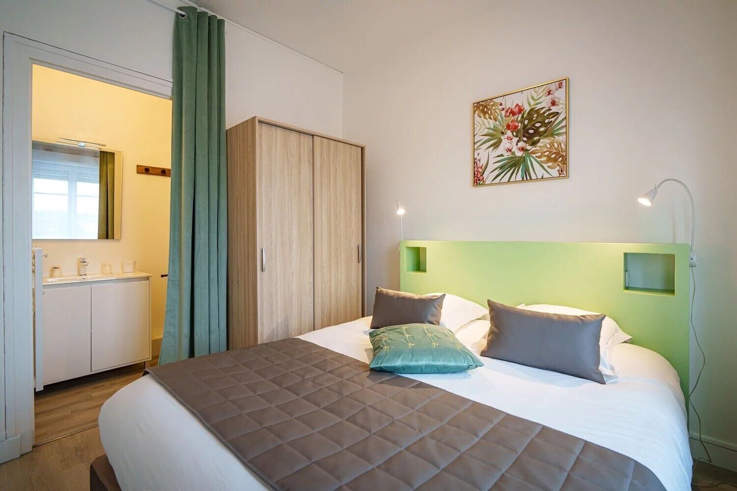 A hotel room with a bed, a green headboard, a bedside table, a nightstand, a wall-mounted lamp, a painting, and a wardrobe.