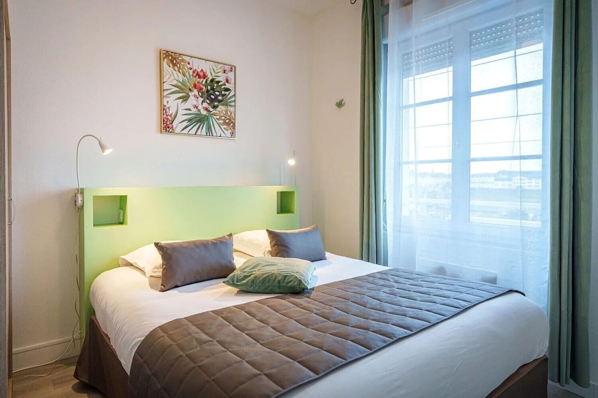 Une chambre d’hôtel avec un lit, une tête de lit verte, une fenêtre donnant sur un beau paysage et une œuvre d’art florale accrochée au mur.