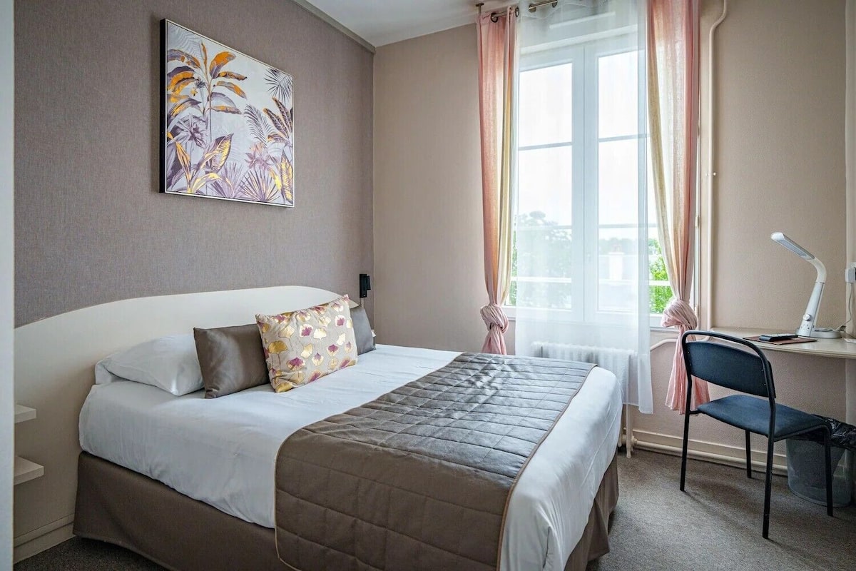 Une chambre d’hôtel avec un lit, un bureau et une chaise, une fenêtre avec des rideaux et un tableau au mur.