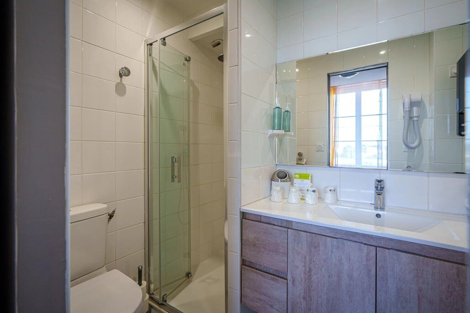 Un baño moderno con ducha envuelta en vidrio, lavamanos blanco con espejo arriba y muebles de madera.