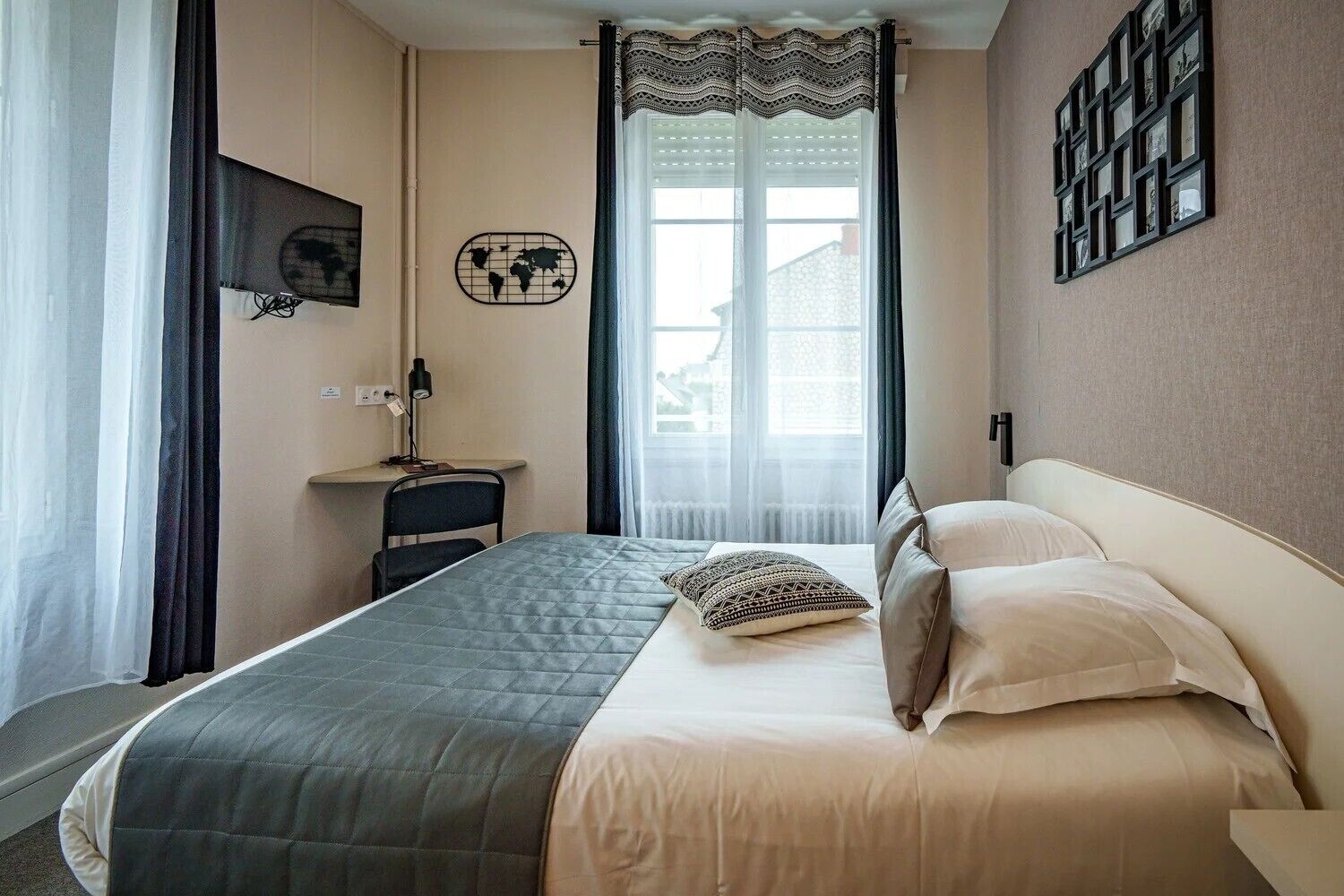 A hotel room with a bed, a desk, a chair, a lamp, a world map decoration, and a window with curtains.