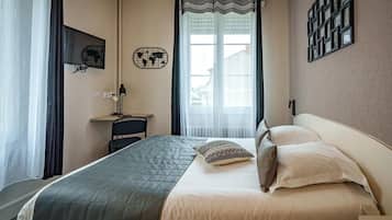 A hotel room with a bed, a desk, a chair, a lamp, a world map decoration, and a window with curtains.