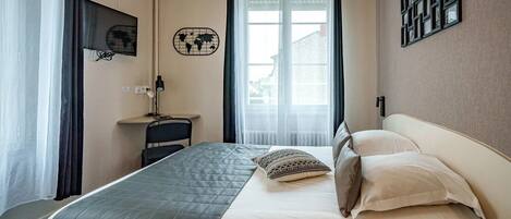A hotel room with a bed, a desk, a chair, a lamp, a world map decoration, and a window with curtains.