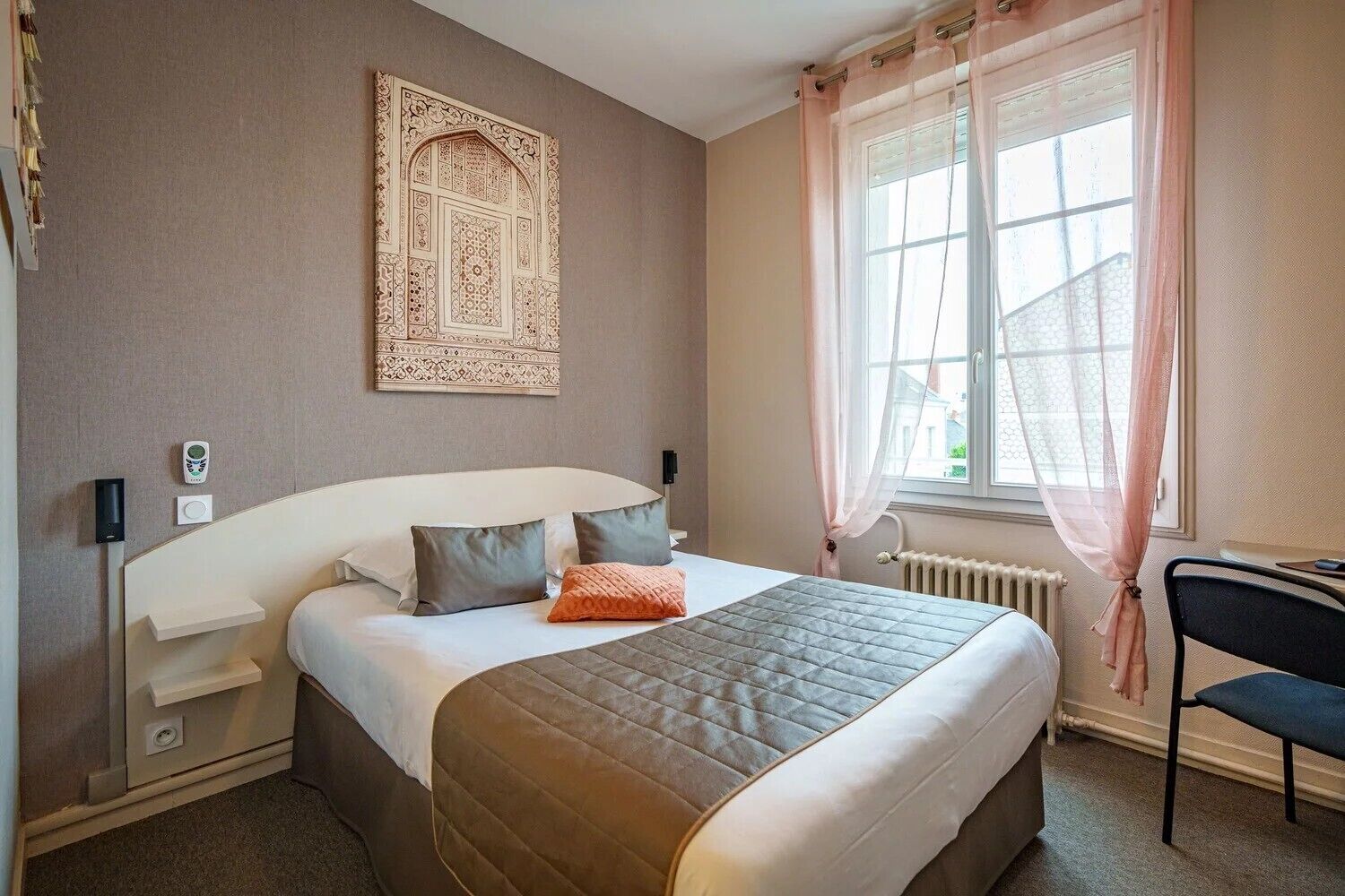 A hotel room with a large bed, a bedside table, a chair, a window with curtains, and a decorative wall piece.