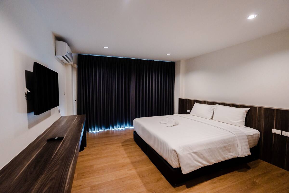 Una habitación de hotel moderna con una cama grande, un televisor de pantalla plana montado en la pared y un fondo de cortina oscura.