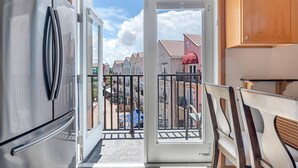 Une cuisine moderne dotée d’un grand réfrigérateur, d’un meuble en bois et d’une porte-fenêtre coulissante donnant sur un balcon avec vue sur des bâtiments aux couleurs vives et un terrain de jeux.