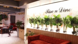 Interior de un restaurante con un letrero 'Rise n Dine', techo de madera, plantas en macetas y un sofá rojo.
