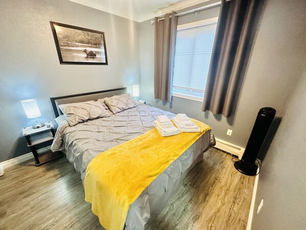 Un dormitorio con cama, mesita de noche con lámpara, ventana con cortinas y un cuadro en la pared.