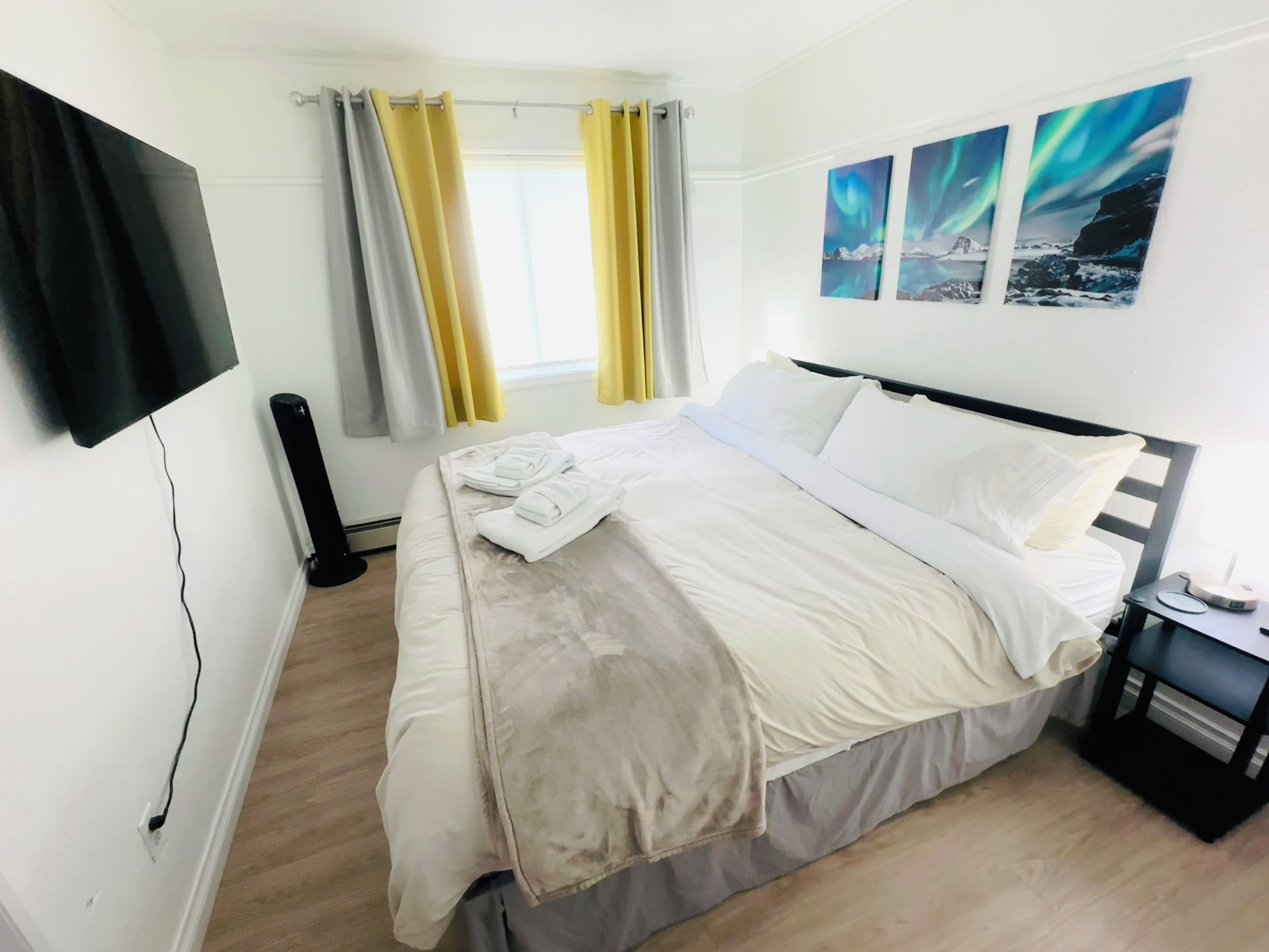 A hotel room with a bed, a flat-screen TV mounted on the wall, a window with curtains, and artwork on the wall.