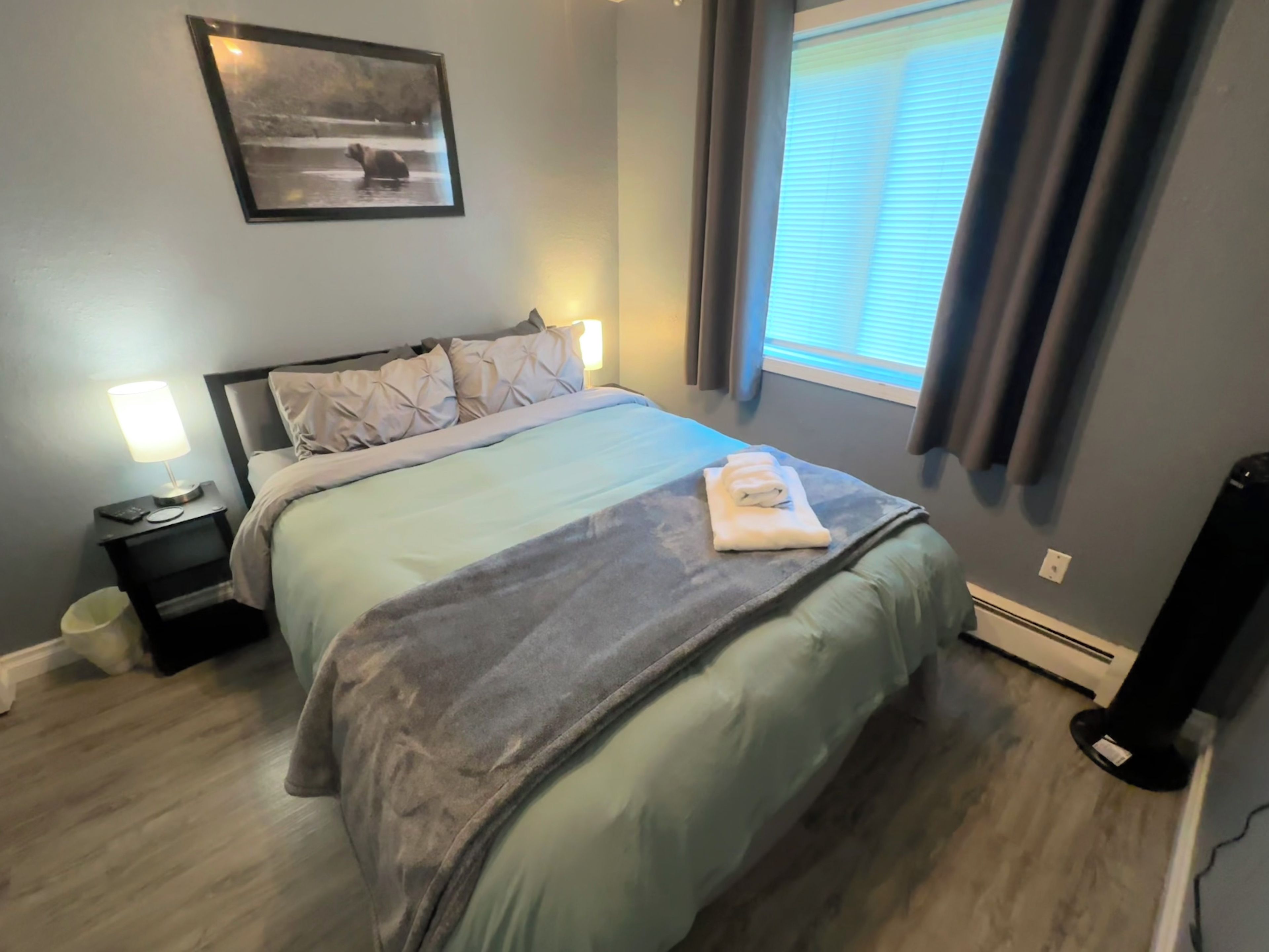 A bedroom with a bed, bedside lamps, a nightstand, a picture on the wall, a window with curtains, and a speaker.