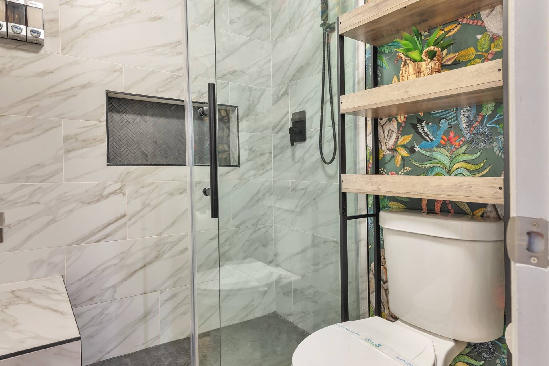 Een badkamer met een glazen douchecabine, een toilet met een kleurrijke muurschildering en een achterwand van marmer met een herringbone-patroon.