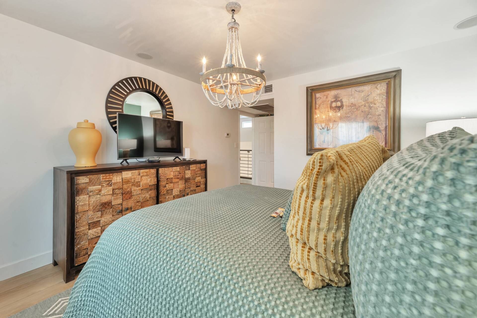 Une chambre à coucher avec un lit, un téléviseur posé sur une commode en bois, un lustre et un tableau accroché au mur.