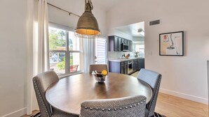 A dining area with a round table, four chairs, a hanging pendant light, and a view of the kitchen and outside.