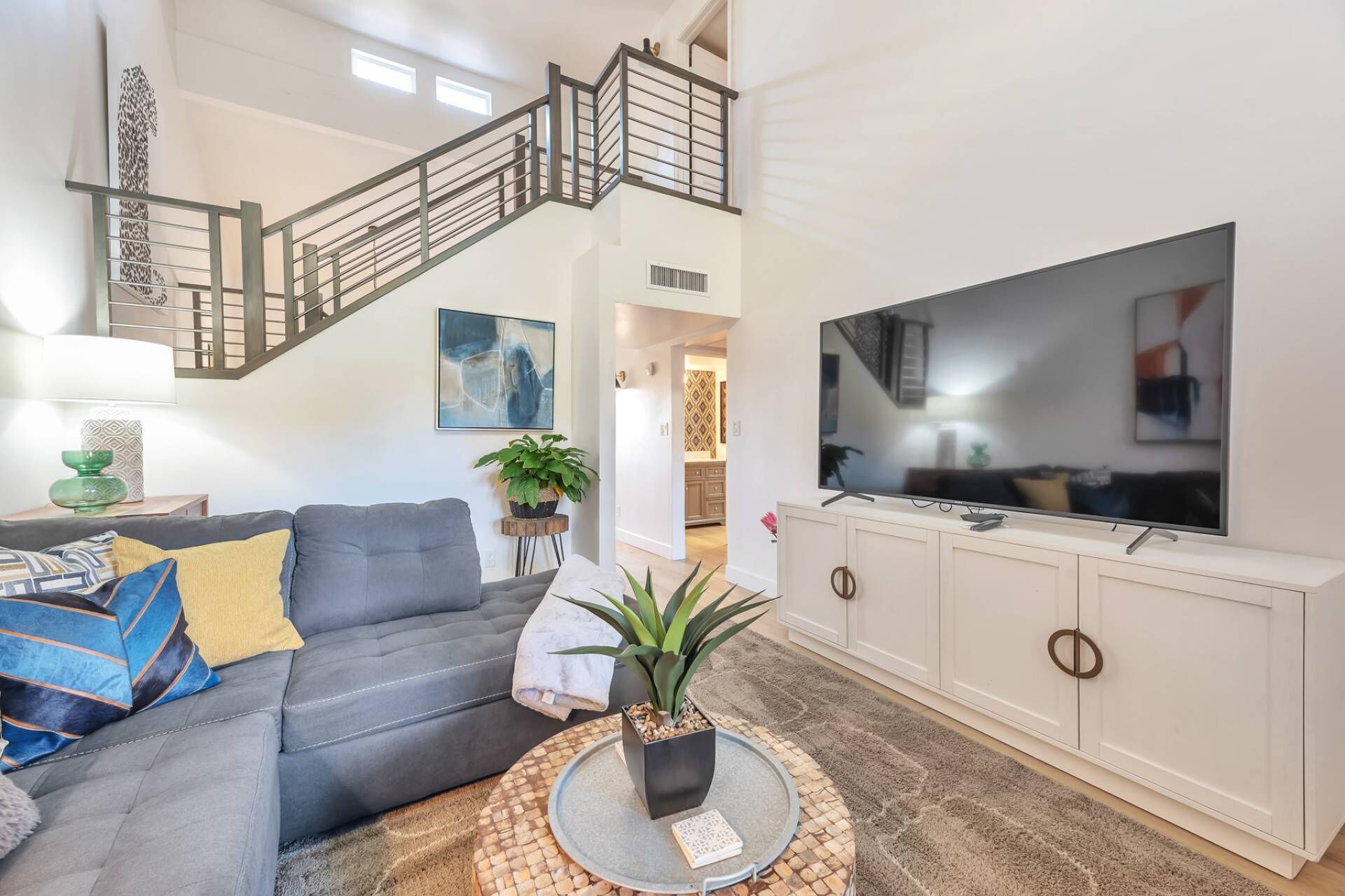 A living room with a grey sofa, a round coffee table with a plant, a TV on a white cabinet, and a staircase with a metal railing.