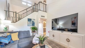 A living room with a grey sofa, a round coffee table with a plant, a TV on a white cabinet, and a staircase with a metal railing.