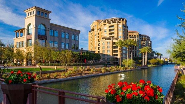 A riverside walkway with a modern building and a multi-story hotel.