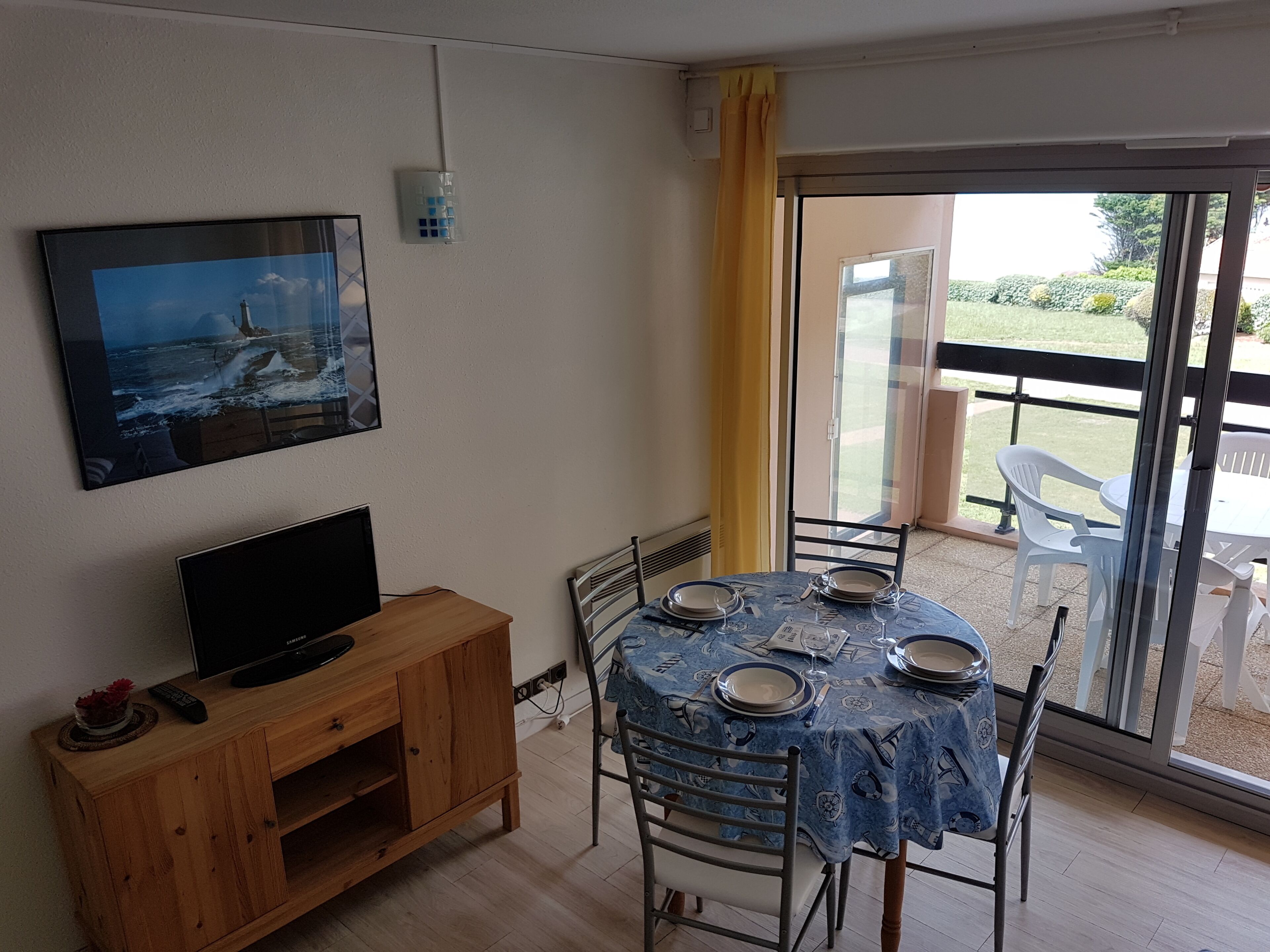 Un comedor con una mesa lista para cuatro personas, un televisor sobre un mueble de madera y vistas al exterior a través de un ventanal.
