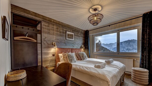 A bedroom with a wooden headboard, a bed with white linens, a desk with a chair, and a view of mountains through a large window. - Appartement Mont Parrain 2627 with Sauna - La Tzoumaz, 4 Vallees (La Tzoumaz)