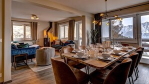 A modern dining area with a long table set for six, surrounded by brown leather chairs. A cozy seating area with a blue sofa and a fireplace is visible in the background. - Appartement Mont Parrain 2627 with Sauna - La Tzoumaz, 4 Vallees (La Tzoumaz)