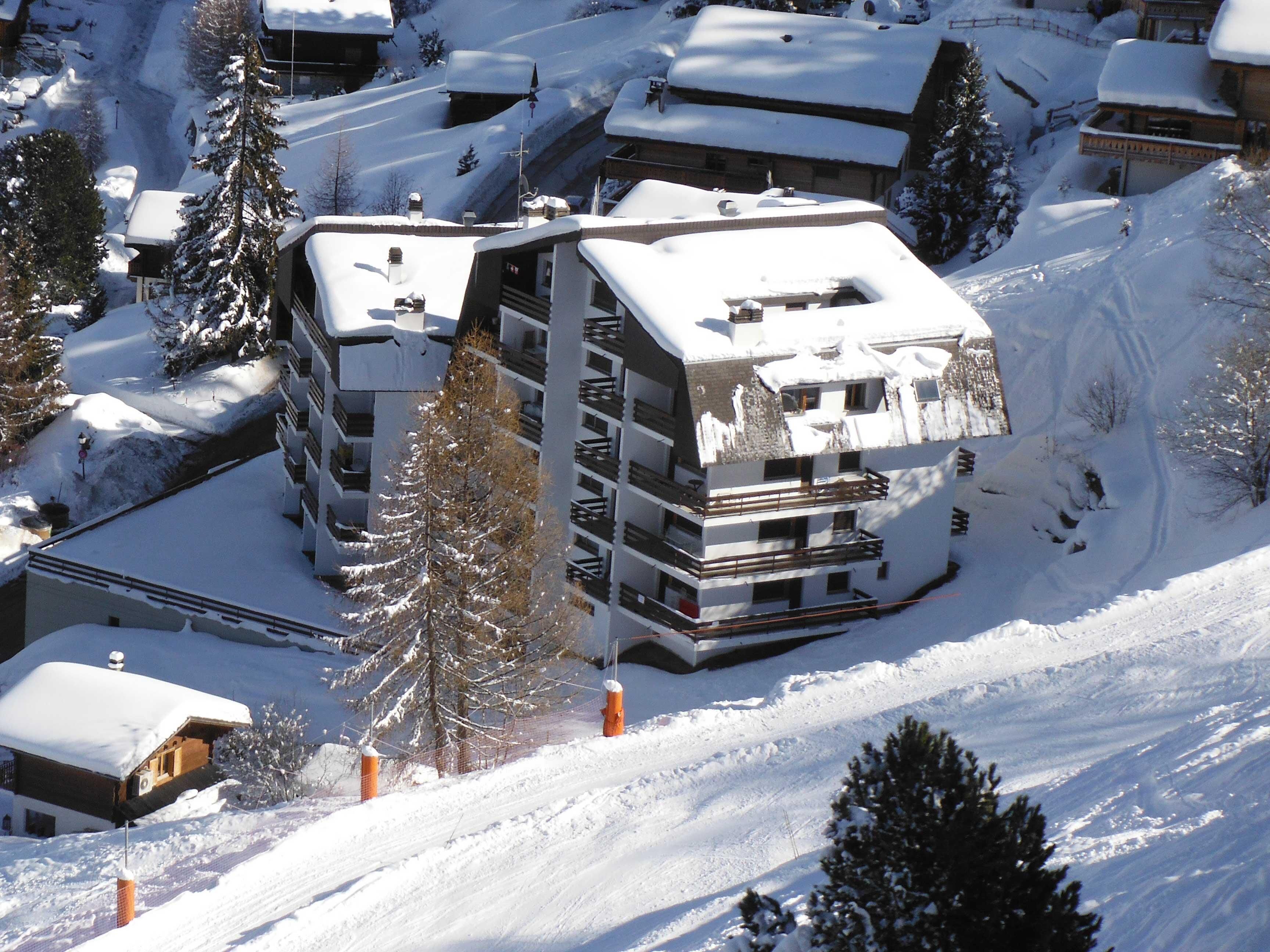 Un edificio de varios pisos con tejado cubierto de nieve, rodeado de árboles nevados y una pista de esquí.