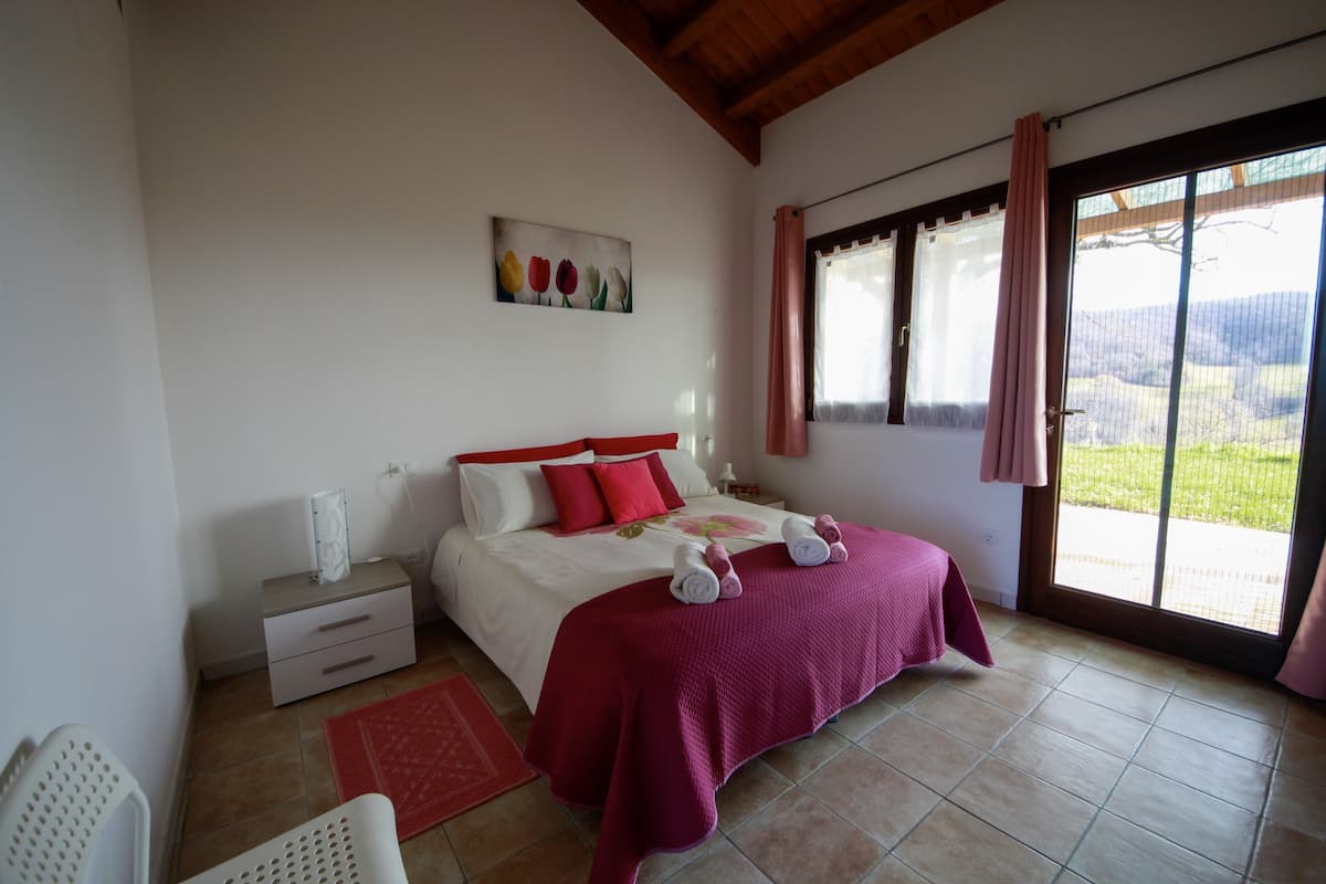 Ein Schlafzimmer mit einem Bett, einem Nachttisch, einem Sessel, einem Fenster mit Vorhängen und einem Bild an der Wand.
