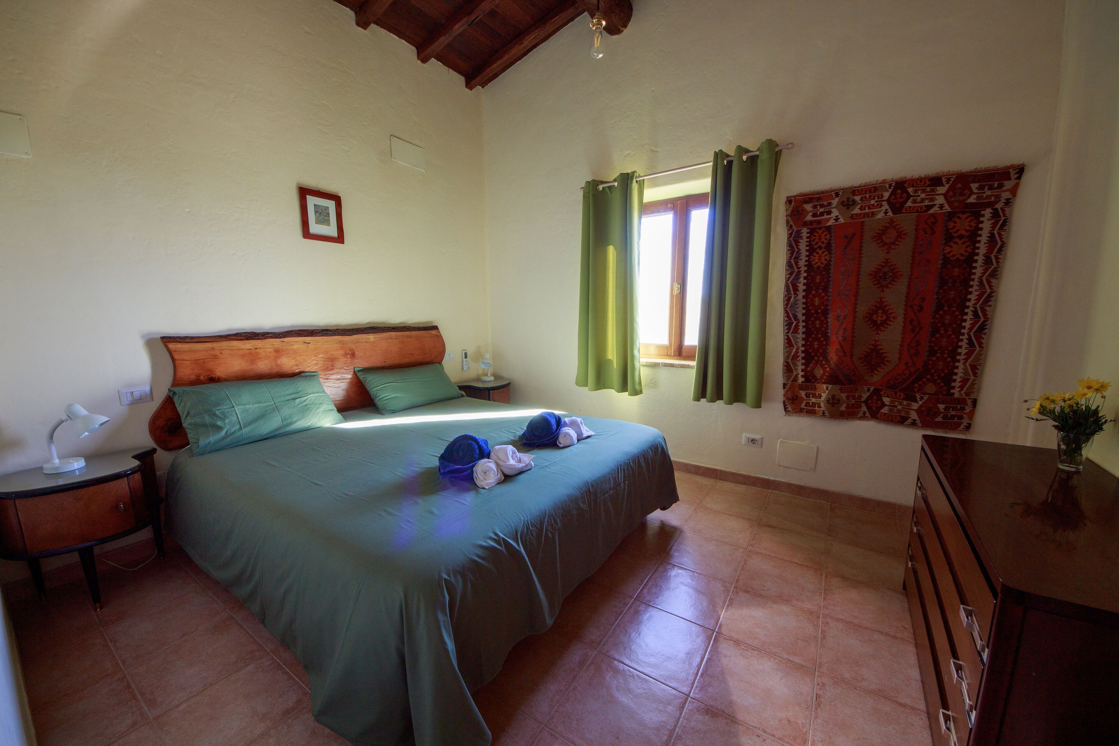 A bedroom with a bed, a nightstand, a dresser, a window with curtains, and a wall-mounted tapestry.