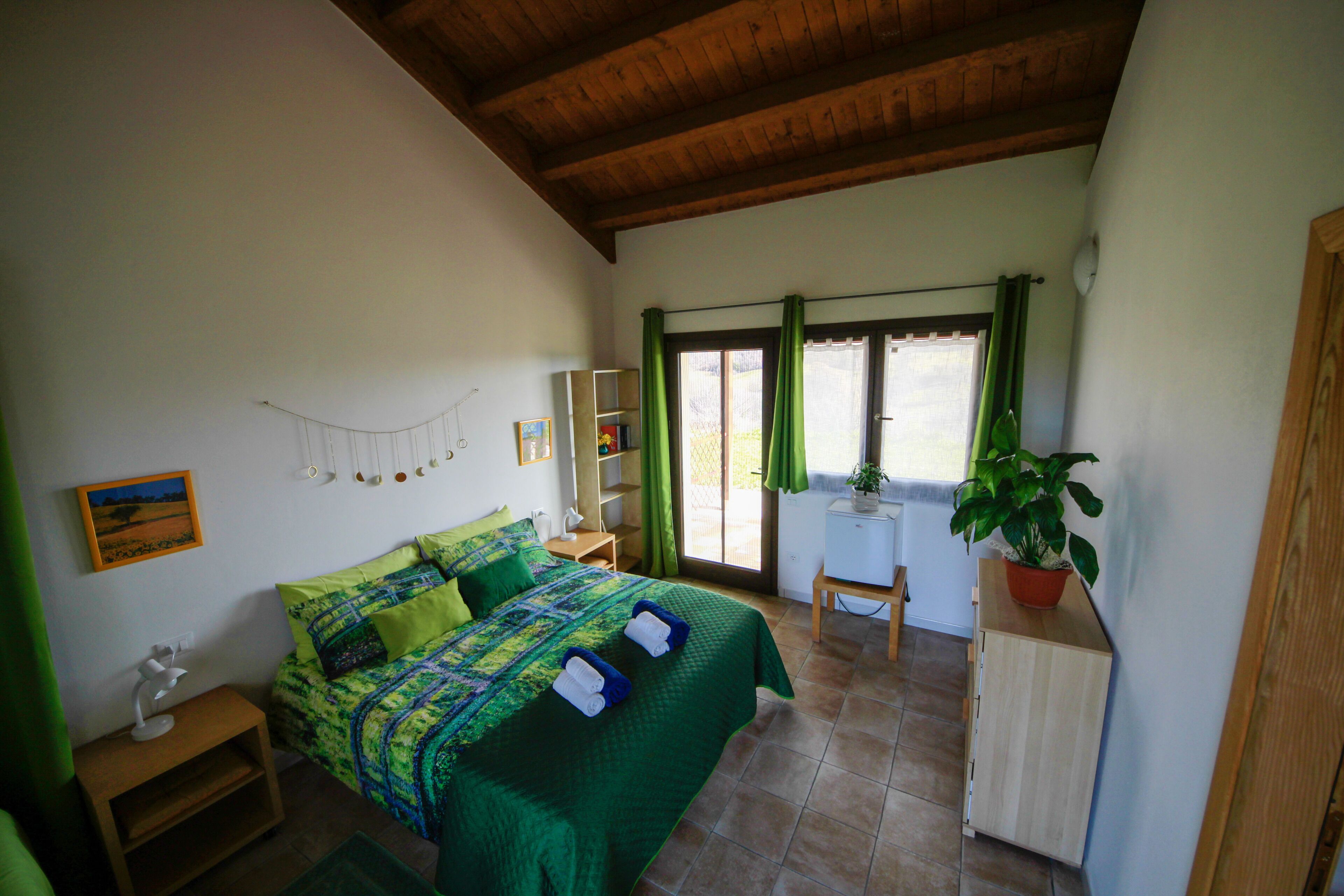 Ein Schlafzimmer mit einem Bett, einem Schreibtisch, einem Stuhl, einer Topfpflanze und einem Fenster mit Vorhängen.