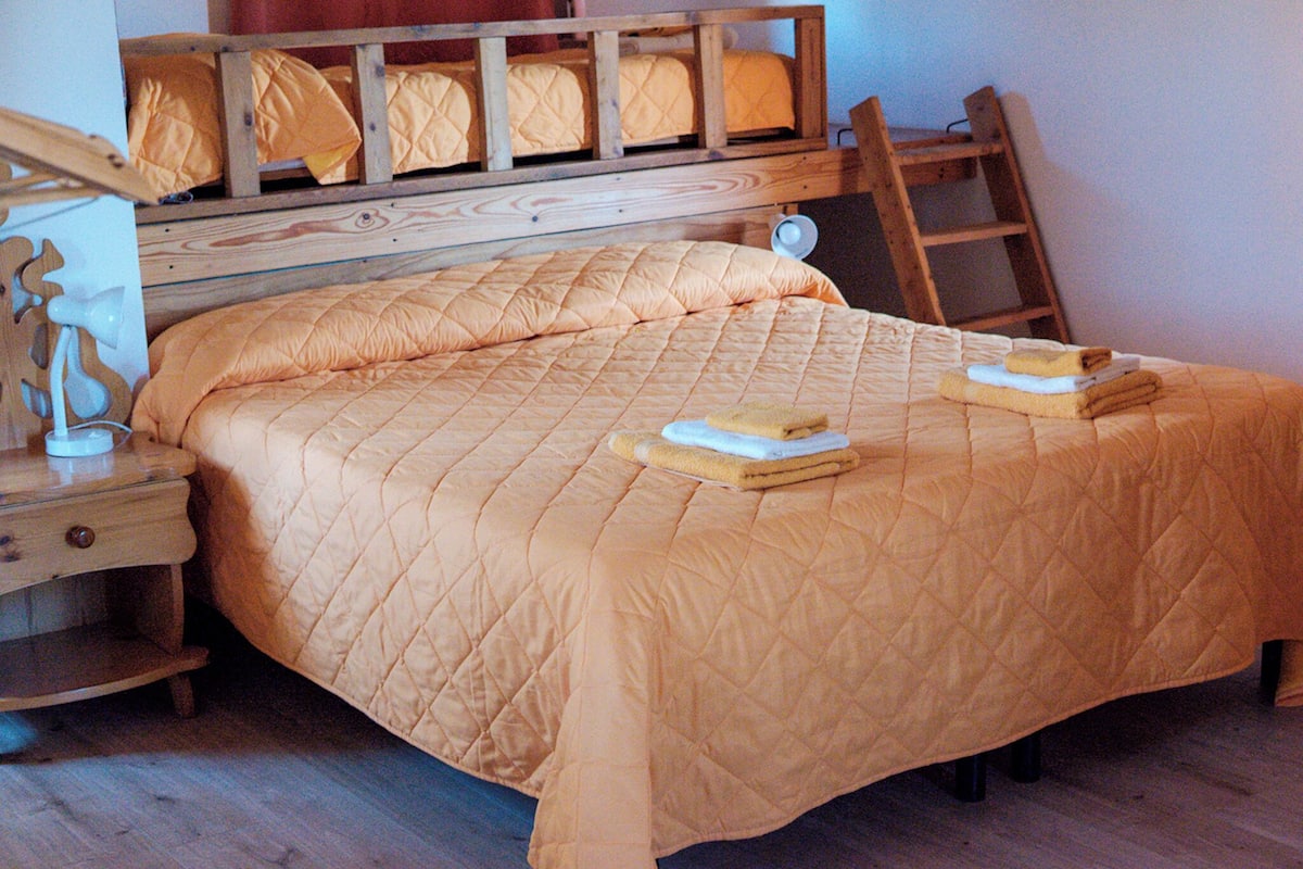 Ein Schlafzimmer mit einem Bettgestell aus Holz, einer orangefarbenen gesteppten Bettdecke, zwei gefalteten Handtüchern auf dem Bett, einem Nachttisch mit einer Lampe und einer Holzleiter, die an der Wand lehnt.