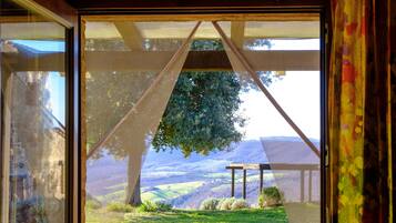 Vista a través de una ventana con una pérgola de madera y un árbol, sobre un paisaje con colinas y praderas.