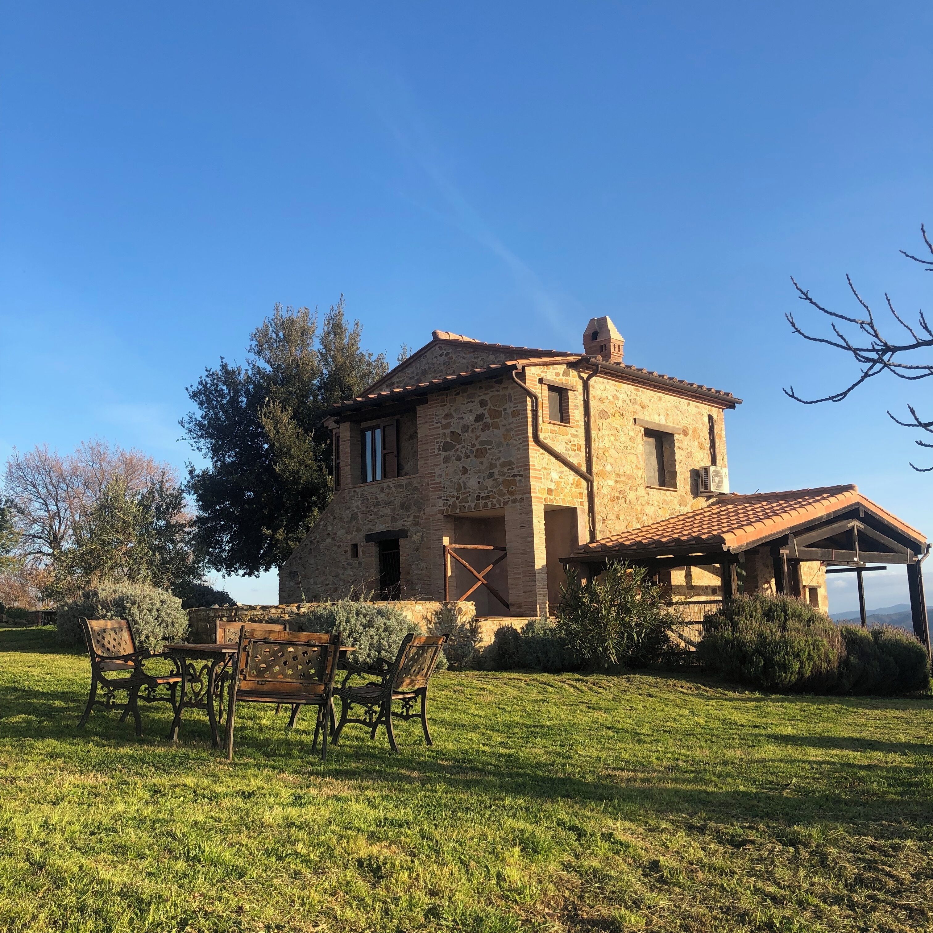 A stone building with a tiled roof, a wooden pergola, and a grassy lawn with a table and chairs.