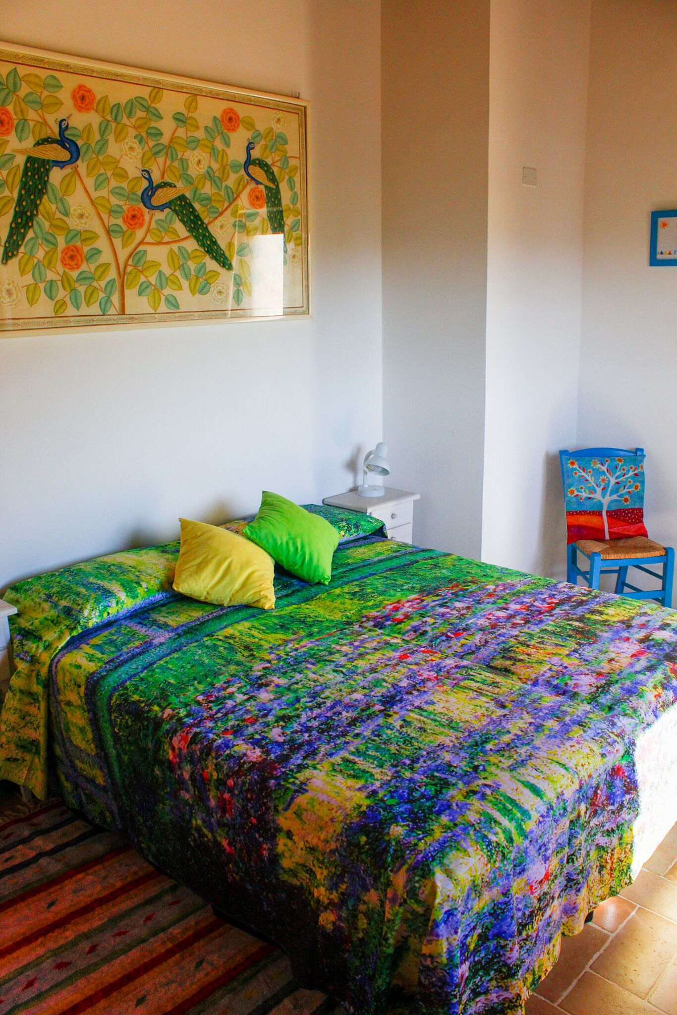 A bedroom with a colorful bedspread, a painting of peacocks, a chair, and a nightstand with a lamp.