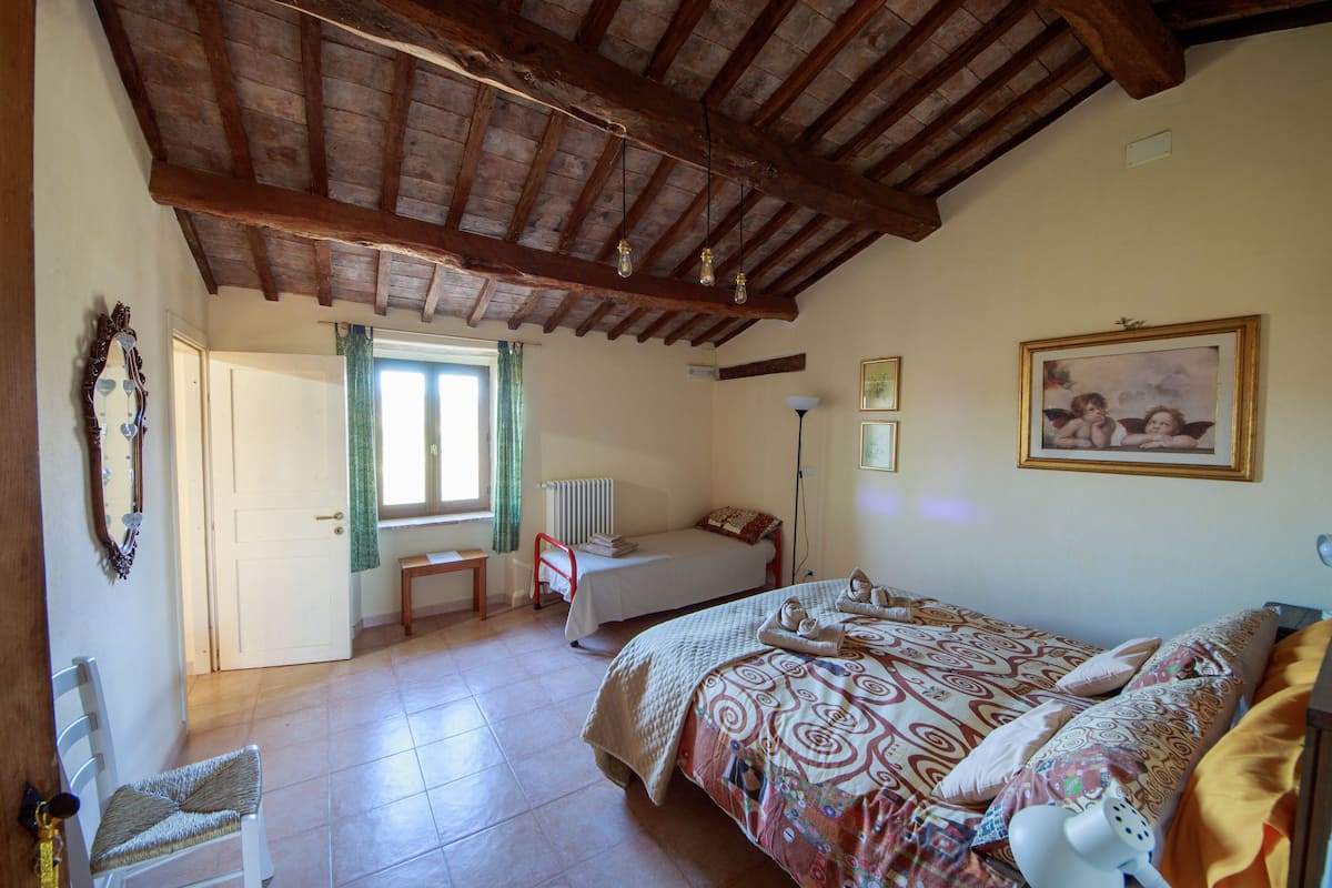 Ein Schlafzimmer mit Holzboden, einem Bett mit gemusterten Betttextilien, einem Sessel, einem Nachttisch und einem Fenster mit Vorhängen.