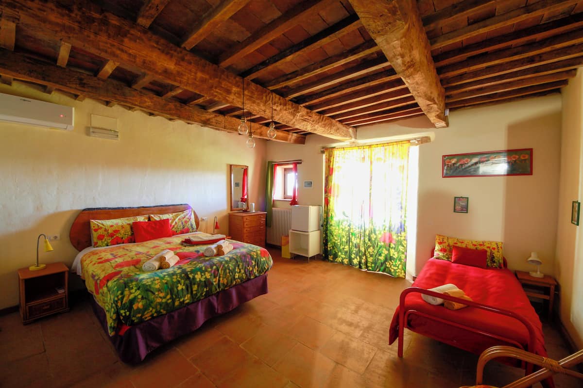 Ein Schlafzimmer mit Holzboden, einem Bett mit Blumenbettwäsche, einem Sessel, einem Nachttisch, einer Kommode und einem Fenster mit Vorhängen.