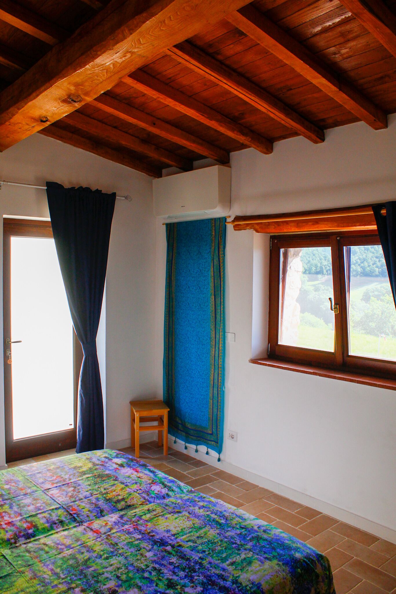A bedroom with a wooden ceiling, a bed with a colorful quilt, a window with curtains, and a small wooden stool.
