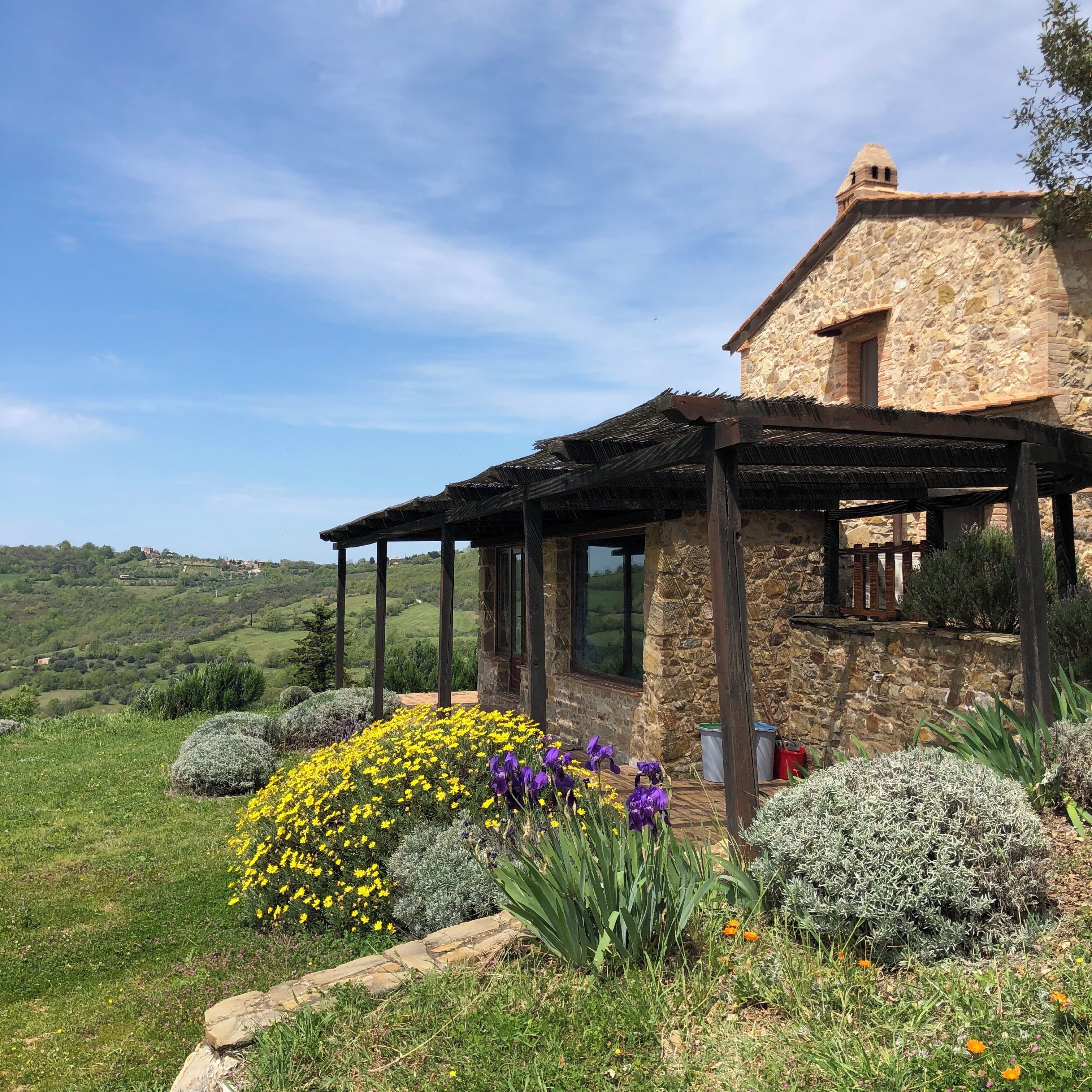 A stone house with a covered patio, surrounded by a garden with yellow and purple flowers.