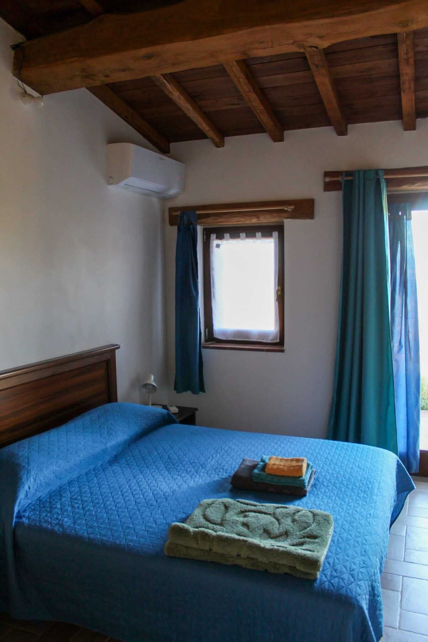 A bedroom with a wooden ceiling, a bed with blue bedding, a window with curtains, and a small table with a lamp.