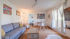 Un salon moderne avec un canapé, une table basse et un coin repas avec des chaises. Il y a un téléviseur sur le côté droit et des œuvres d’art accrochées aux murs.