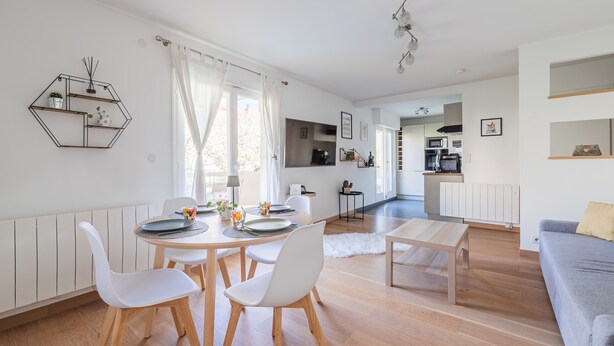 Un salon moderne avec un coin repas, un plancher en bois, des murs blancs et une cuisine à l’arrière-plan.