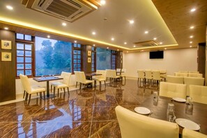 A spacious conference room with a polished marble floor, multiple tables, and chairs, large windows with a view of trees, and a flat-screen TV mounted on the wall.