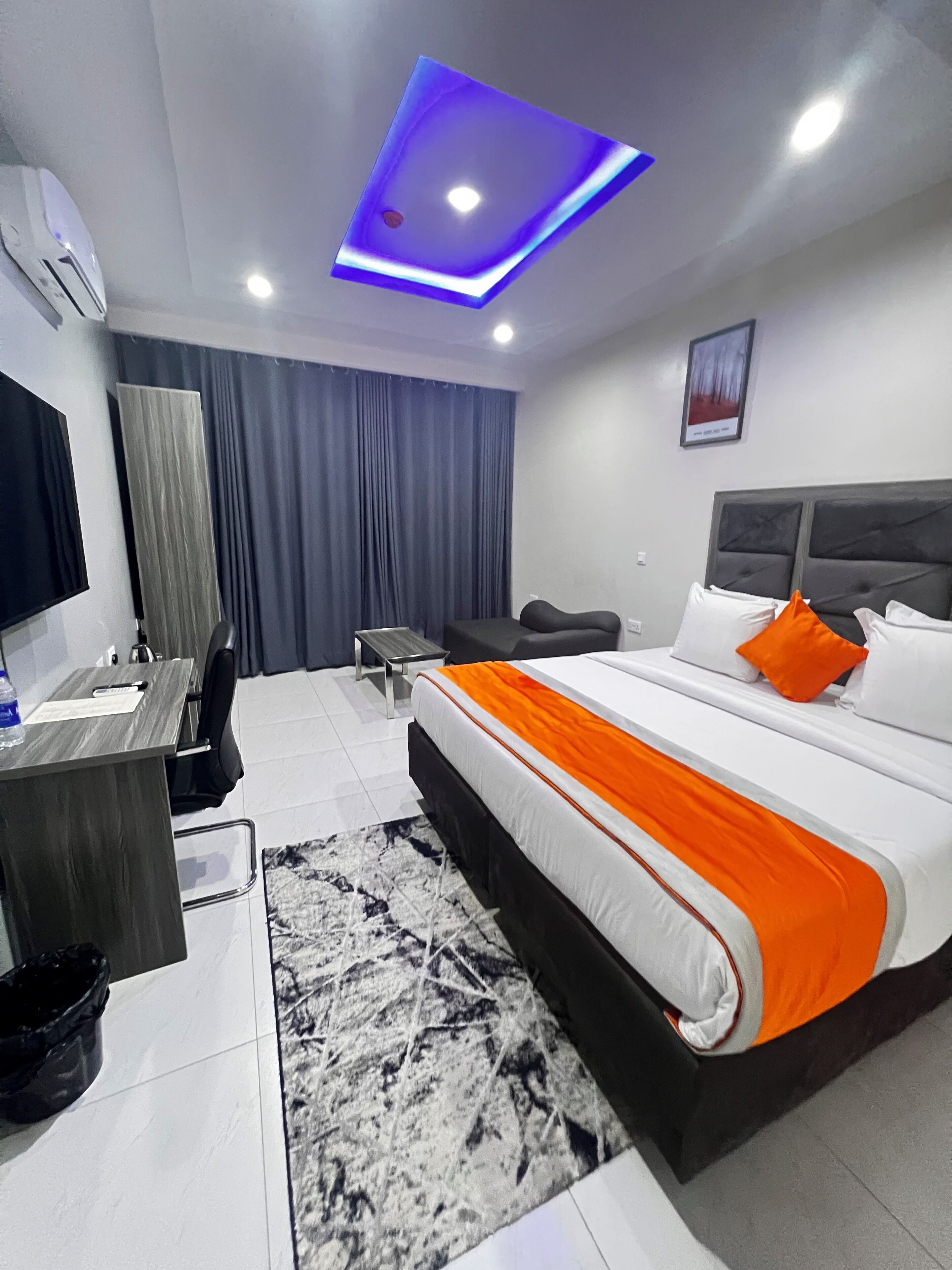 A modern hotel room with a large bed, a desk, a chair, a TV, and a ceiling light.